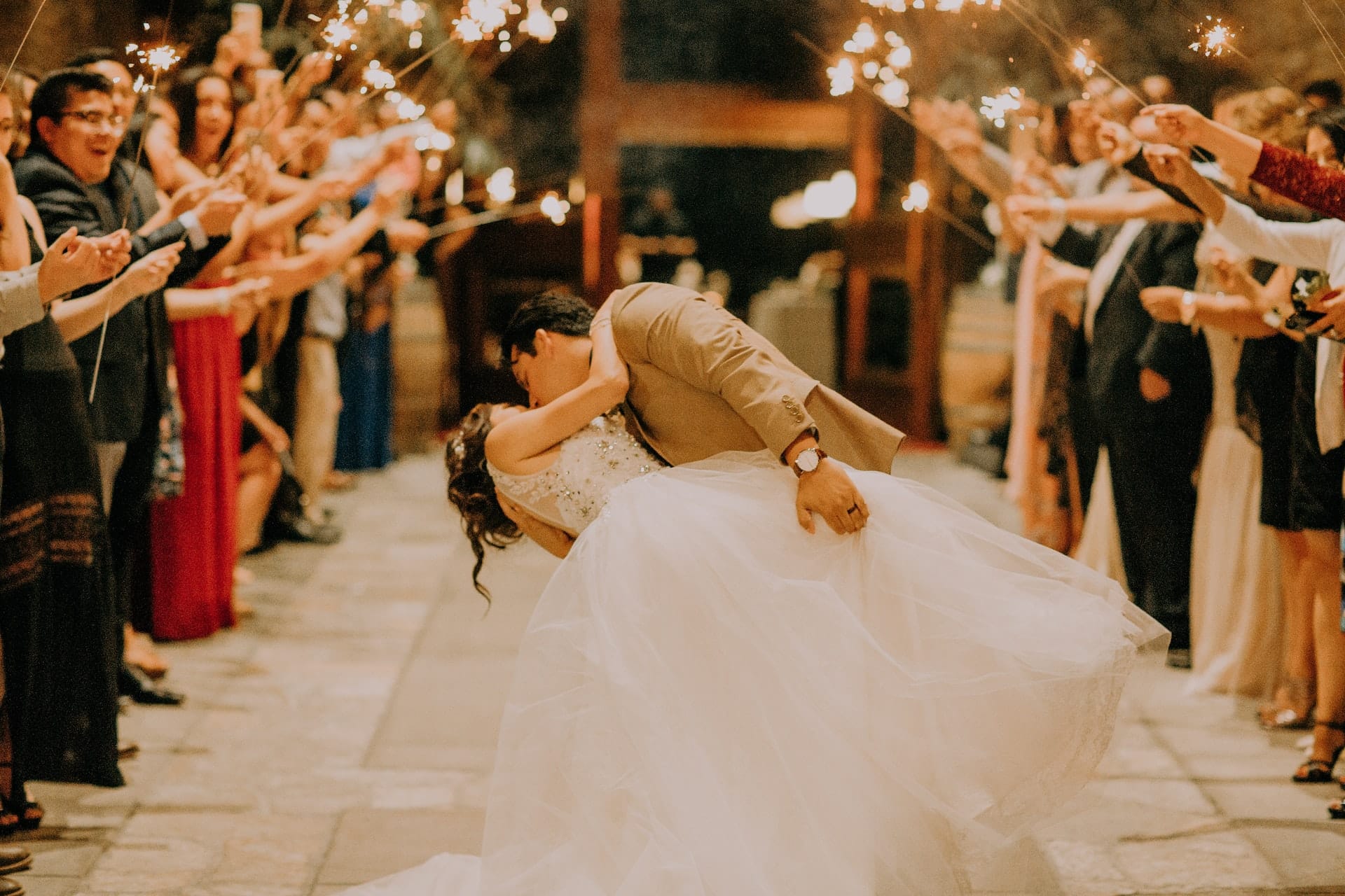Bride and groom in a dip, kissing while guests hold sparklers around them in two lines surrounding a path